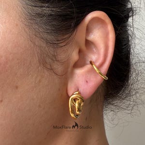 May include: Close-up of a gold-colored ear cuff and a pair of gold earrings. The earrings have a knotted design. The ear cuff is a simple, curved band. The jewelry is worn on a person's ear.