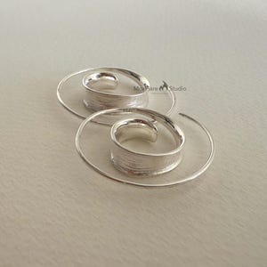 May include: A pair of silver spiral earrings. Each earring has a textured, cylindrical centre and a delicate, spiralling wire design. The earrings are displayed on a light-coloured surface, showcasing their minimalist and modern aesthetic.