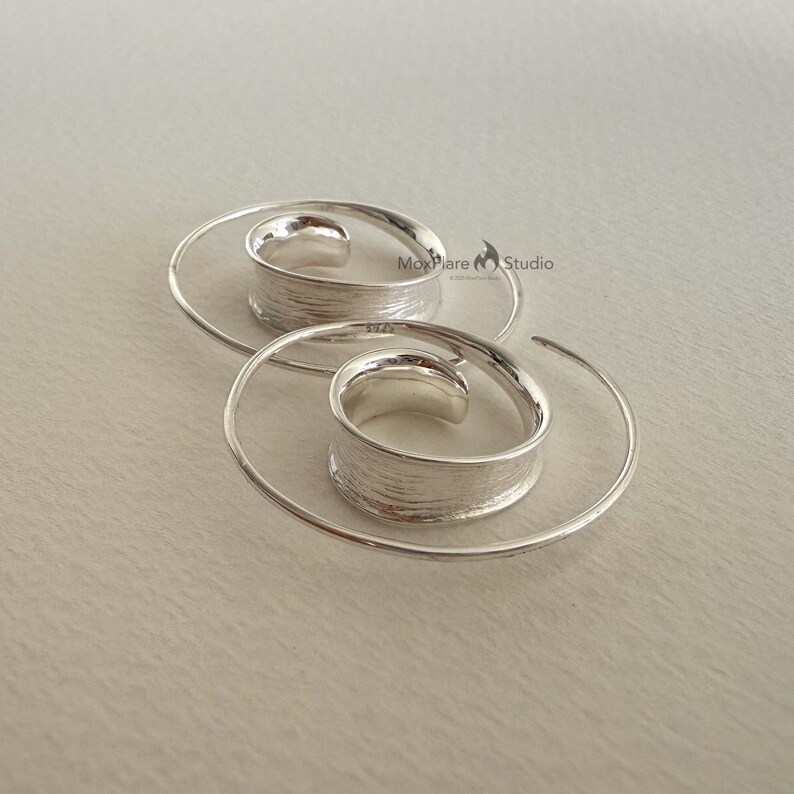 May include: A pair of silver spiral earrings. Each earring has a textured, cylindrical centre and a delicate, spiralling wire design. The earrings are displayed on a light-coloured surface, showcasing their minimalist and modern aesthetic.