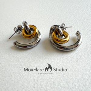 May include: A pair of silver-tone and gold-tone hoop earrings. Each earring features a curved, polished silver hoop with a smaller gold-tone ring interlocked at the top. The earrings have a post-back closure. The brand name "MoxFlare Studio" is at the bottom.