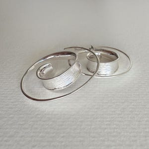 May include: A pair of silver spiral earrings. Each earring has a textured, curved silver element that spirals outwards, connected to a circular band. The earrings are displayed on a light background.