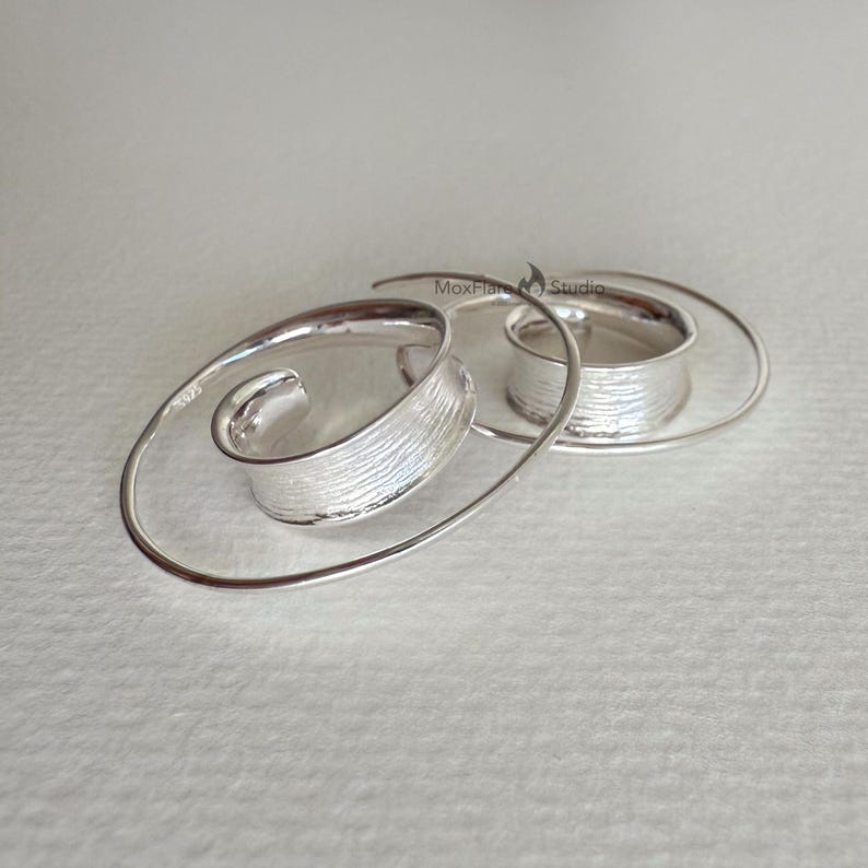 May include: A pair of silver spiral earrings. Each earring has a textured, curved silver element that spirals outwards, connected to a circular band. The earrings are displayed on a light background.