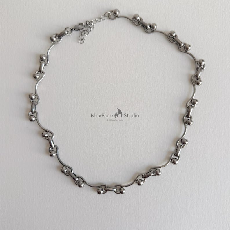 May include: A silver-toned necklace with a unique design. The necklace features a series of interconnected, curved metal segments, each adorned with small, spherical accents. The design is symmetrical and the necklace has an adjustable chain.