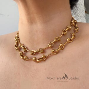 Może przedstawiać: Złoty naszyjnik o podwójnym splocie. Naszyjnik składa się z serii małych, okrągłych koralików połączonych zakrzywionymi, wydłużonymi ogniwami. Naszyjnik jest noszony wokół szyi, prezentując swoje skomplikowane detale i ciepły kolor.