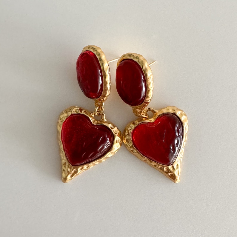 May include: Gold heart-shaped earrings with red glass stones. The earrings have a textured gold finish.