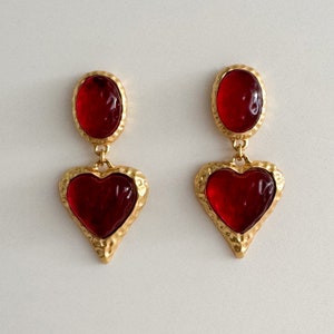 May include: A pair of gold tone earrings with red heart-shaped glass stones. The earrings have a textured gold finish and are dangling.