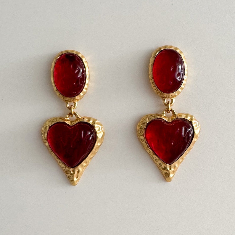 May include: A pair of gold tone earrings with red heart-shaped glass stones. The earrings have a textured gold finish and are dangling.