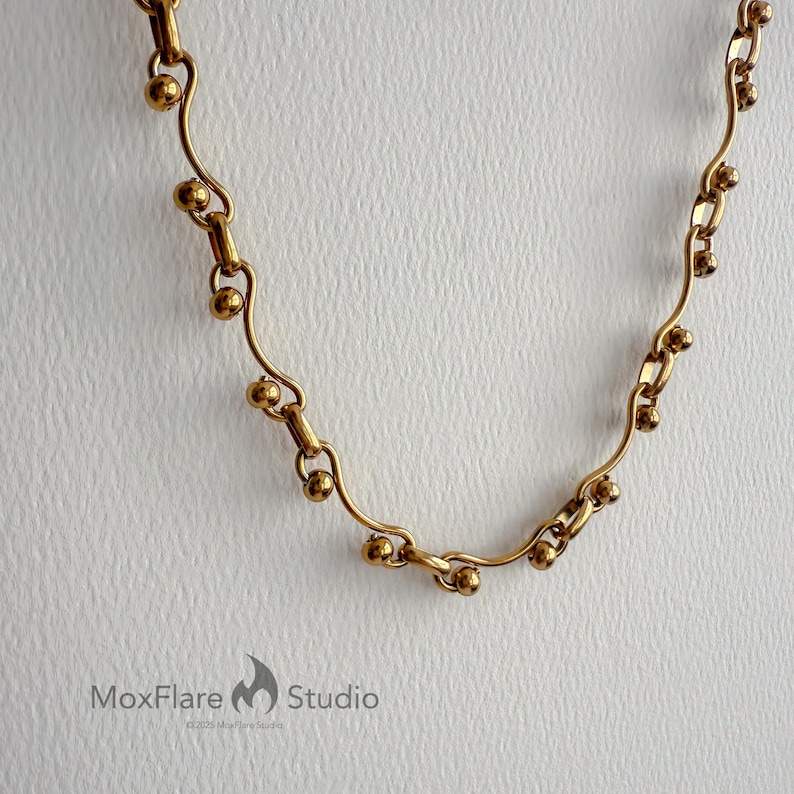 May include: A gold-colored necklace with a unique design. The necklace features a series of small, round beads interspersed along a delicate, curved chain. The design is symmetrical and elegant, with a vintage aesthetic. The MoxFlare Studio logo is visible in the bottom left corner.