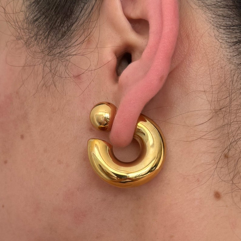 May include: Gold hoop earrings with a ball on the top. The earrings are worn in the earlobe.