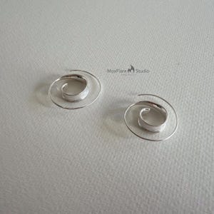 May include: Two silver spiral earrings. Each earring has a coiled design with a textured centre and a delicate, spiralling wire extending outwards. The earrings are displayed on a light grey surface.