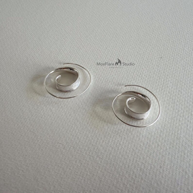 May include: Two silver spiral earrings. Each earring has a coiled design with a textured centre and a delicate, spiralling wire extending outwards. The earrings are displayed on a light grey surface.