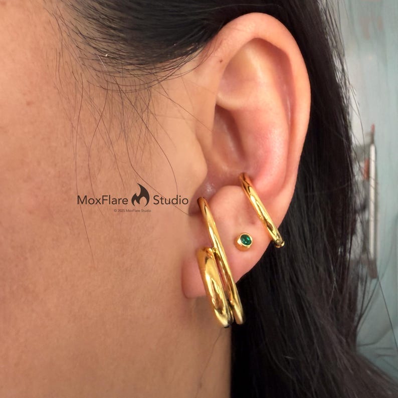 May include: Gold-colored earrings and ear cuffs. The earrings have a double-hoop design, and the ear cuffs curve around the ear. A small, round, green gemstone stud earring is also visible. The image is of a person's ear.