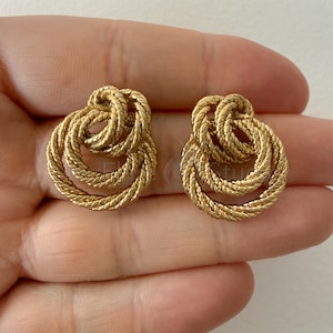 May include: A pair of gold-toned earrings featuring a textured, twisted rope design. Each earring is composed of two interlocking circular loops, creating a layered, knotted appearance with a metallic finish and a consistent, braided surface pattern.