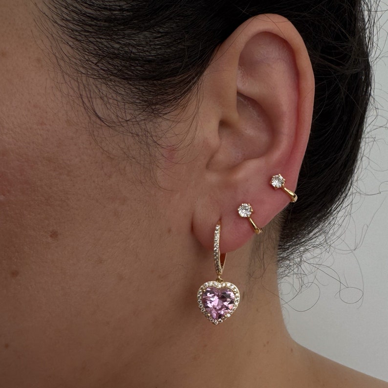 May include: Gold hoop earrings with a pink heart charm and clear crystal accents. The earrings are worn in the earlobe and helix piercings.