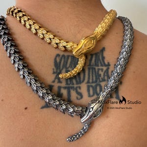 May include: Two snake-shaped necklaces. One is gold-colored, the other is silver-colored. The necklaces are made of a chain-like material, with the snake heads and tails detailed. The gold necklace is positioned above the silver one.