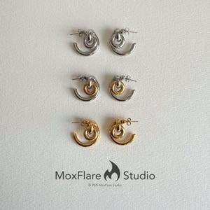 May include: Three pairs of earrings in silver, silver and gold, and gold. Each pair features a curved, open design with a circular element at the top. The earrings are displayed on a white surface. The brand name "MoxFlare Studio" is at the bottom.
