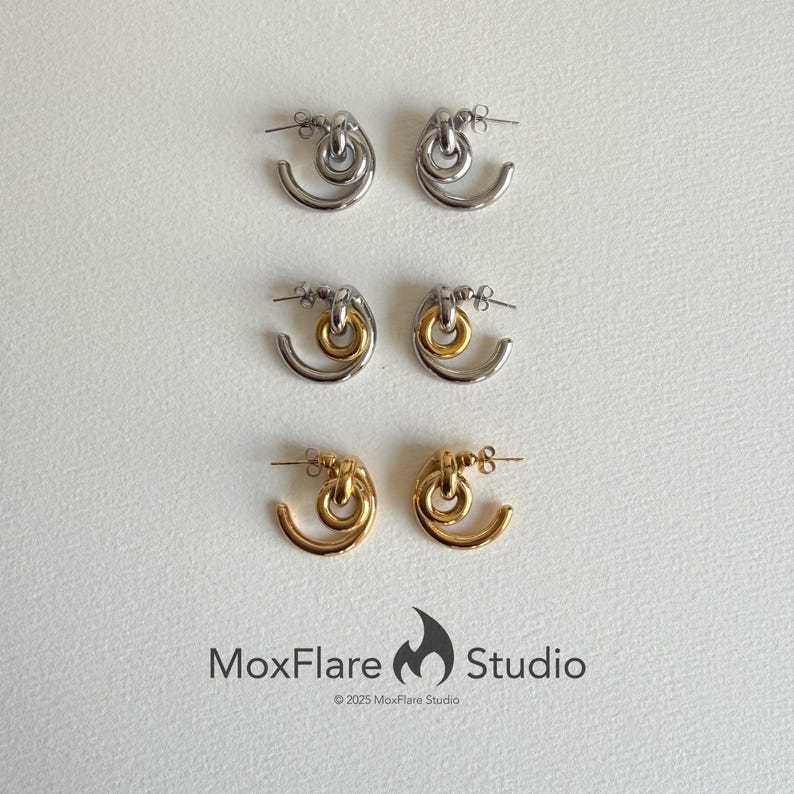 May include: Three pairs of earrings in silver, silver and gold, and gold. Each pair features a curved, open design with a circular element at the top. The earrings are displayed on a white surface. The brand name "MoxFlare Studio" is at the bottom.