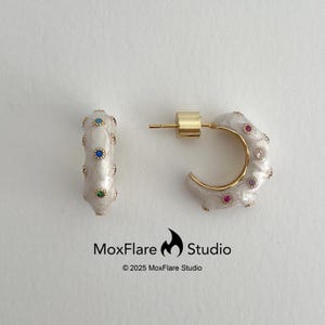 Peut inclure: Boucles d'oreilles créoles dorées avec un émail blanc et des pierres précieuses colorées. Ces boucles d'oreilles, de MoxFlare Studio, ont une tige dorée et une forme de croissant.