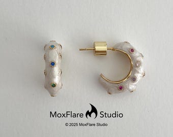 May include: A pair of hoop earrings with a gold-tone metal base. The earrings have a white enamel finish with colorful gemstones and a gold post. The earrings are from MoxFlare Studio.