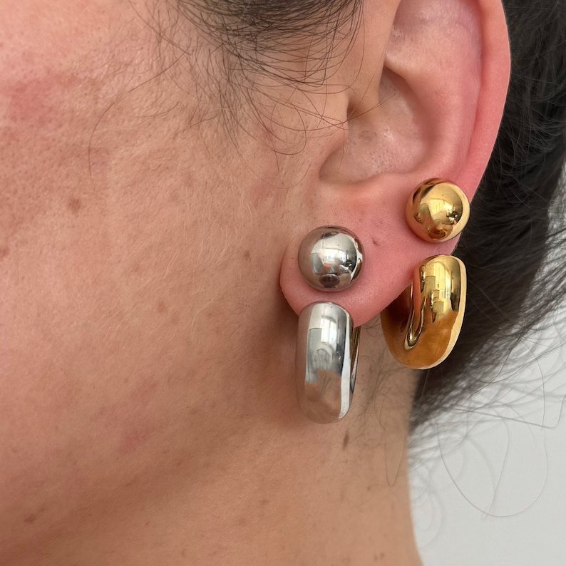 May include: A pair of hoop earrings with a silver and gold finish. The earrings are in the shape of a hoop with a ball at the top. The earrings are worn in the earlobe.