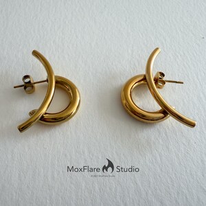 May include: A pair of gold-colored stud earrings. Each earring features a curved, elongated design that loops around a circular base. The earrings have a polished finish and a post-and-clutch closure.
