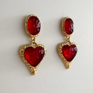 May include: A pair of gold tone dangle earrings with red heart-shaped glass stones. The earrings have a textured gold finish.