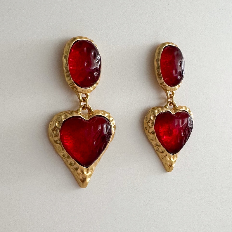 May include: A pair of gold tone dangle earrings with red heart-shaped glass stones. The earrings have a textured gold finish.