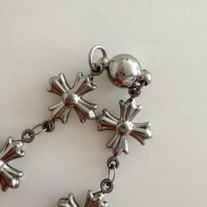 May include: A silver chain bracelet with a magnetic clasp. The chain is made up of small, interconnected cross-shaped links.