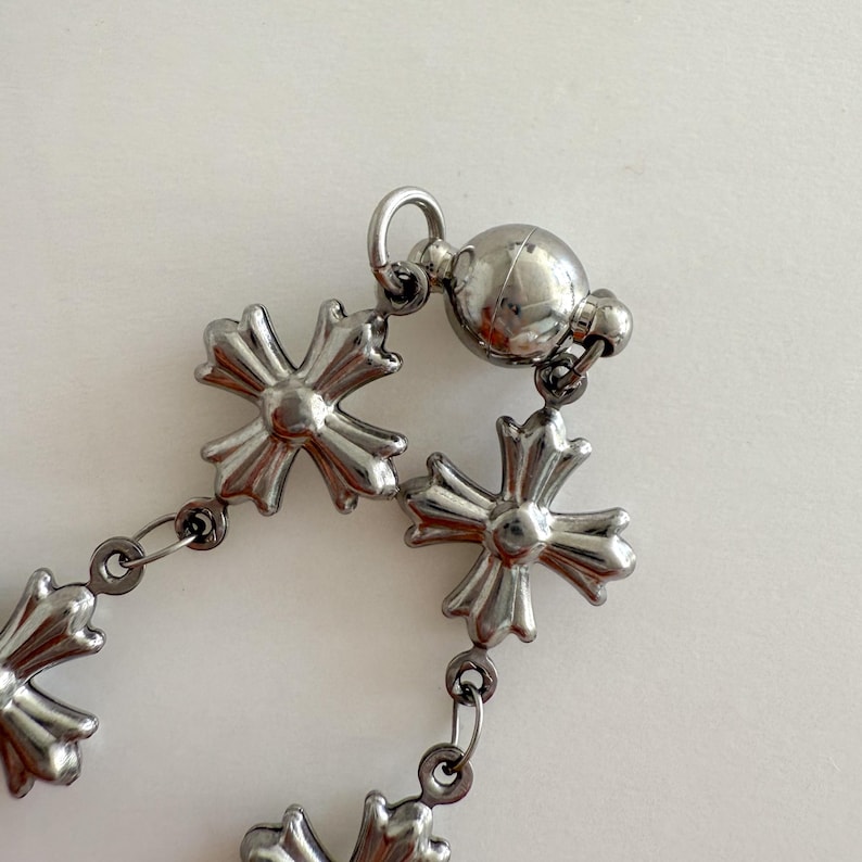 May include: A silver chain bracelet with a magnetic clasp. The chain is made up of small, interconnected cross-shaped links.