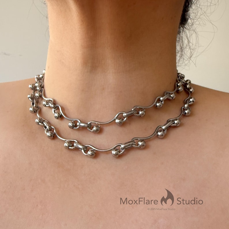 May include: A silver-toned necklace with a double-strand design. The necklace features a series of small, round beads and curved links, creating a unique and modern aesthetic. The necklace is worn around the neck.