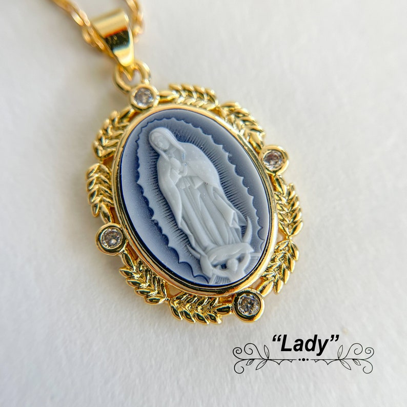 May include: Gold pendant necklace featuring a blue and white cameo of the Virgin Mary. The cameo is surrounded by a gold leaf design and clear rhinestones. "Lady" is written below the cameo.