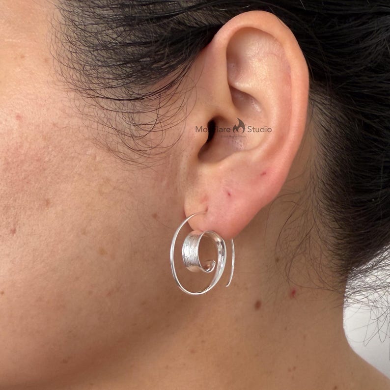 May include: A close-up of a silver spiral earring on an ear. The earring features a coiled design with a textured centre and a smooth, curved outer edge. The earring is made of a shiny, metallic material.