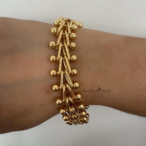 May include: Gold-toned bracelet featuring a unique design of interconnected links and small, round beads. The bracelet is shown on a wrist, showcasing its intricate pattern and polished finish. The MoxFlare Studio logo is visible in the bottom right corner.