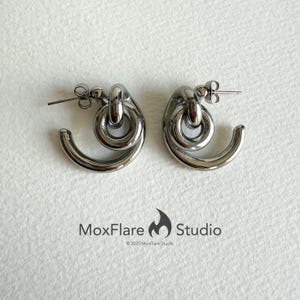May include: A pair of silver-toned hoop earrings. Each earring features a curved, semi-circular design with a loop and a post for pierced ears. The earrings are displayed on a white surface. The text "MoxFlare Studio" and copyright information are visible.