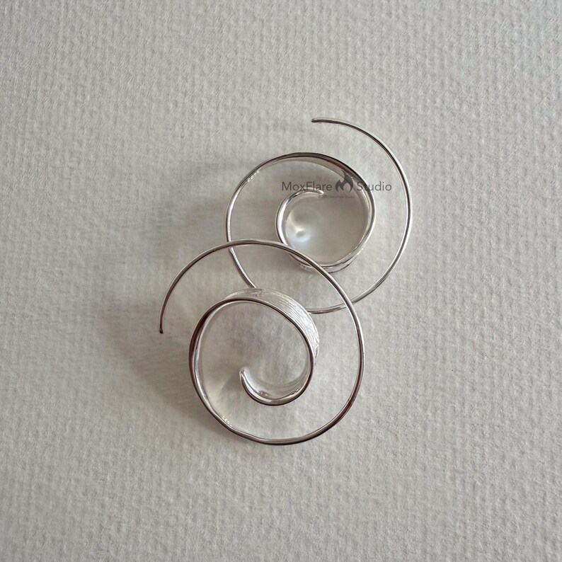 May include: Two silver spiral earrings. Each earring has a coiled design that tapers to a point. The earrings are displayed on a light grey surface. The earrings are from MoxFlare Studio.