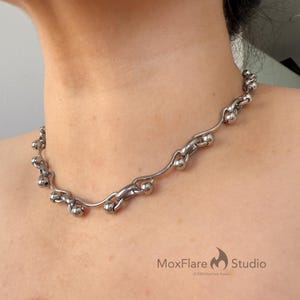 May include: A silver-toned necklace with a unique design. The necklace features a series of interconnected, curved metal elements and spherical beads. The MoxFlare Studio logo is visible in the bottom right corner.