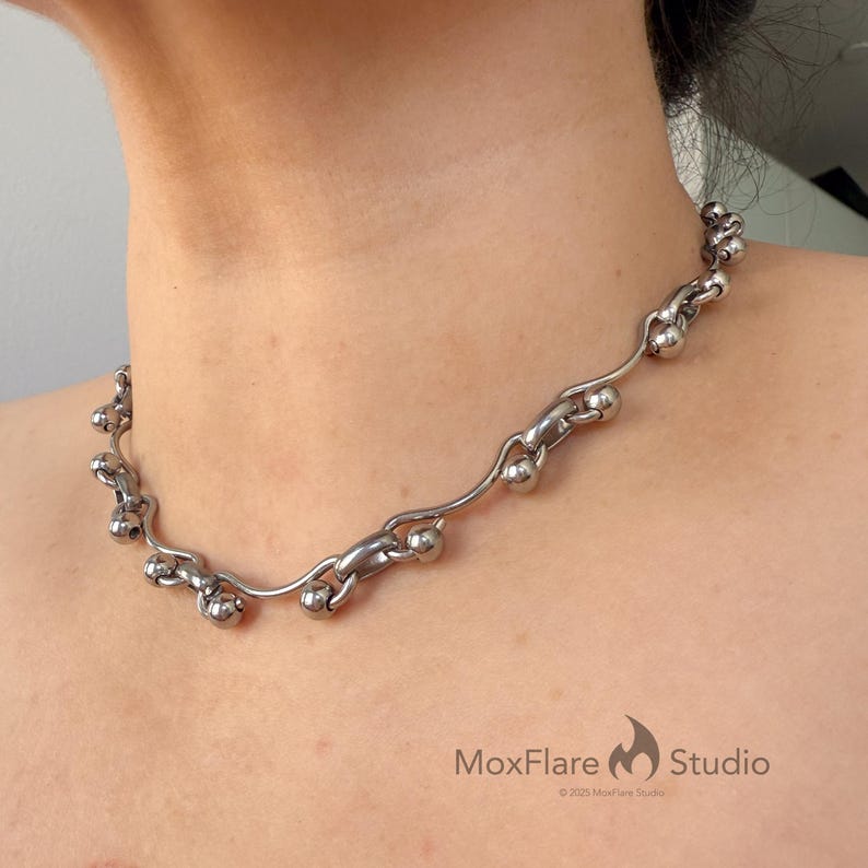 May include: A silver-toned necklace with a unique design. The necklace features a series of interconnected, curved metal elements and spherical beads. The MoxFlare Studio logo is visible in the bottom right corner.