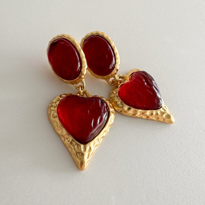 May include: A pair of gold tone earrings with red glass heart-shaped charms. The hearts have a textured gold frame.