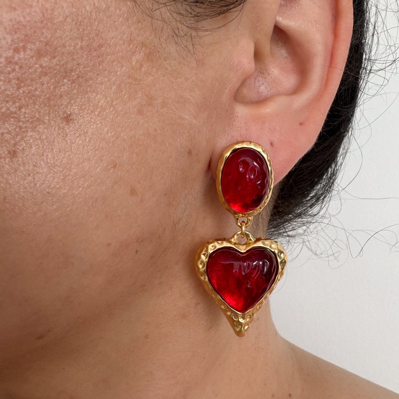 May include: Gold tone dangle earrings with red heart-shaped glass stones. The earrings have a textured gold finish.