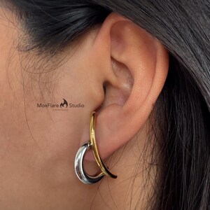 May include: A two-tone earring with a silver teardrop shape and a gold curved accent. The earring is worn on the ear, with the MoxFlare Studio logo visible in the background.
