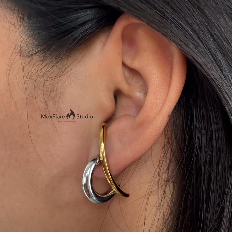 May include: A two-tone earring with a silver teardrop shape and a gold curved accent. The earring is worn on the ear, with the MoxFlare Studio logo visible in the background.