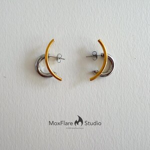 May include: A pair of earrings with a gold-colored curved bar over a silver-colored hoop. The earrings are displayed on a white surface. The earrings are a modern, minimalist design.