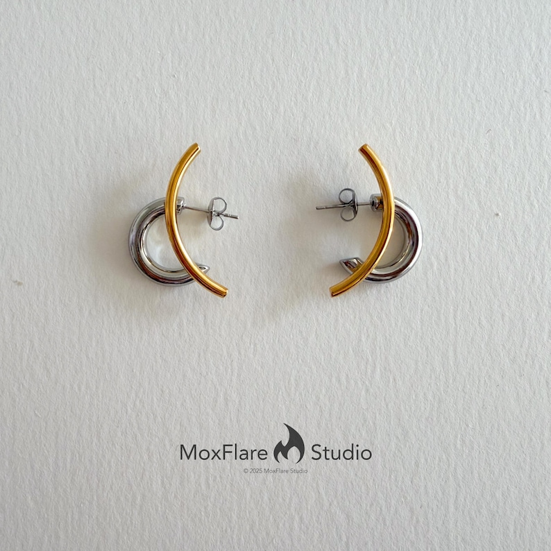 May include: A pair of earrings with a gold-colored curved bar over a silver-colored hoop. The earrings are displayed on a white surface. The earrings are a modern, minimalist design.