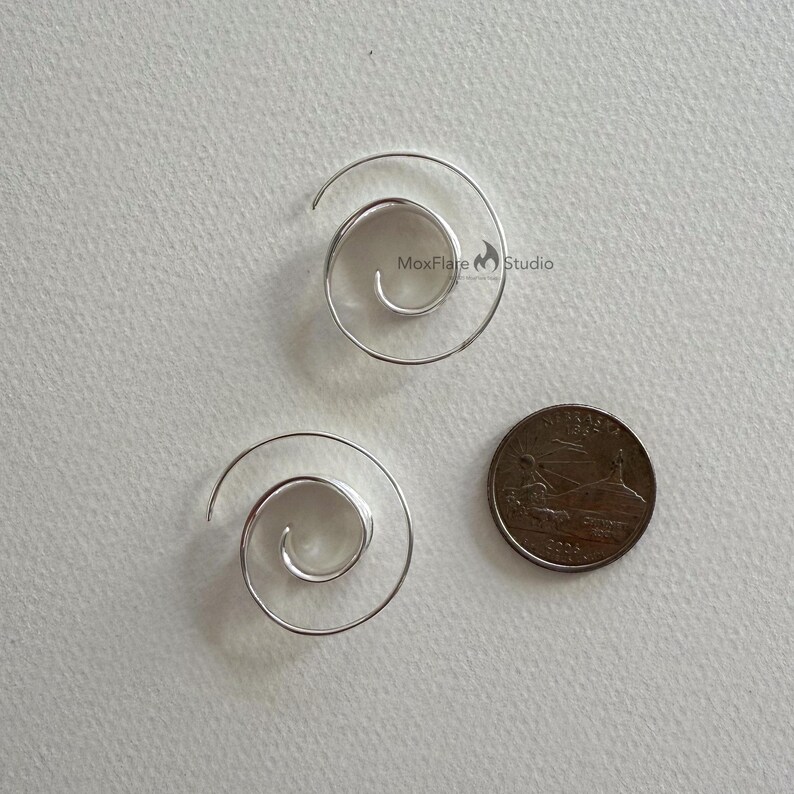May include: Two silver spiral earrings next to a coin. The earrings are made of thin, curved metal, forming a spiral shape. The coin is a UK coin, showing a detailed design.