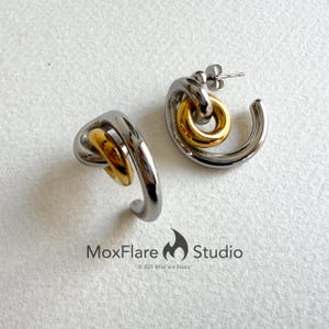 May include: A pair of modern hoop earrings. Each earring features a silver-toned, curved design with a gold-toned circular accent. The earrings are displayed on a white surface. The MoxFlare Studio logo is visible.