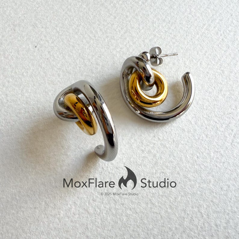 May include: A pair of modern hoop earrings. Each earring features a silver-toned, curved design with a gold-toned circular accent. The earrings are displayed on a white surface. The MoxFlare Studio logo is visible.
