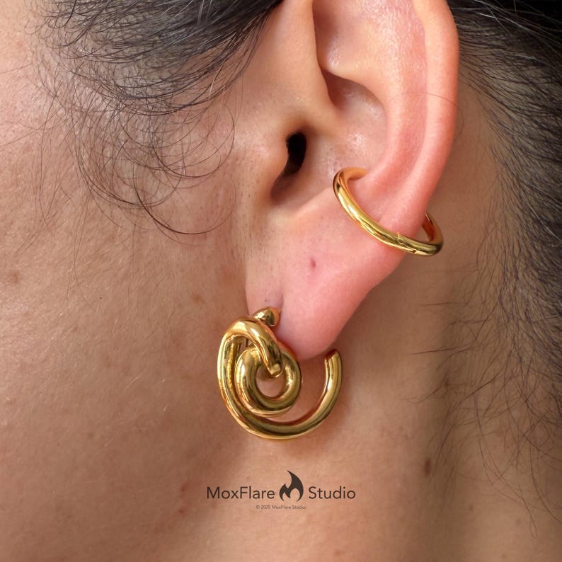 May include: Gold-colored earrings with a knot design and a matching ear cuff. The earrings have a spiral shape and are worn on the earlobe. The ear cuff is a simple band that wraps around the upper ear. The earrings and ear cuff are made of a shiny metal.