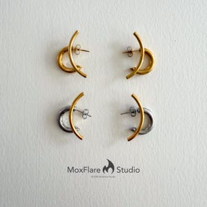 May include: Four pairs of earrings. Two pairs are gold-toned, featuring a curved bar and a circular element. The other two pairs are silver-toned, with a gold-toned curved bar and a silver circular element. The MoxFlare Studio logo is at the bottom.
