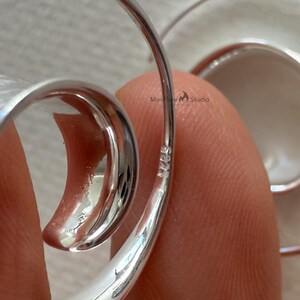 May include: Close-up of a silver earring with a curved, open design. The earring has a smooth, polished finish and a subtle, reflective quality. The metal is stamped with "925" indicating sterling silver.