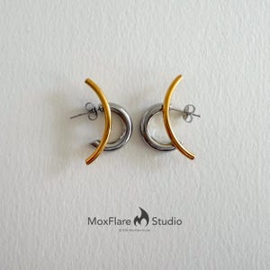 May include: A pair of modern earrings with a gold-colored curved bar accenting a silver-colored hoop. The earrings are displayed on a white surface. The brand name "MoxFlare Studio" is printed in black text.
